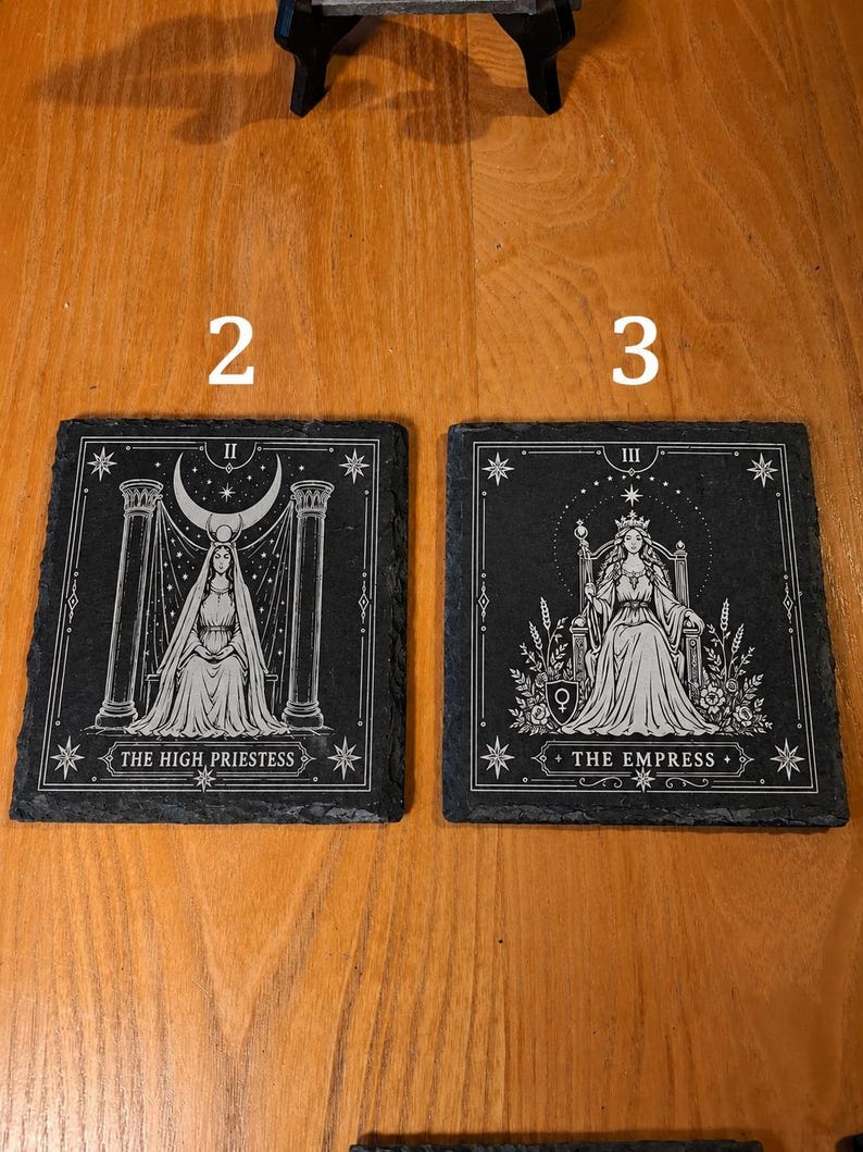 May include: Two square black slate coasters featuring white engraved tarot card designs. The left coaster displays The High Priestess with pillars and a crescent moon. The right coaster shows The Empress seated on a throne with floral details.