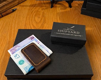 Könnte beinhalten: Ein brauner Leder-Geldscheinclip mit gelben Nähten hält zwei britische Banknoten auf einer schwarzen Schachtel. Eine kleine schwarze Schachtel mit dem Text "SHIPYARD" befindet sich neben dem Geldclip. Ein Schild mit dem Text "SHIPYARD" ist im Hintergrund.
