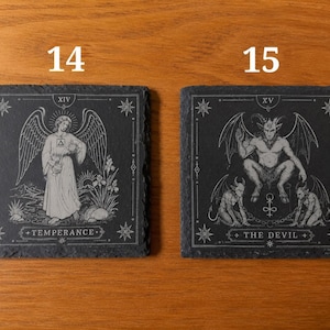 May include: Two square black slate coasters featuring white engraved tarot card designs. The left coaster, labeled 14, depicts Temperance with an angel. The right coaster, labeled 15, depicts The Devil with horned figures. Both have jagged edges.