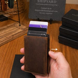 May include: A brown leather cardholder with several cards visible. The cardholder is held in a hand, with a black box in the foreground. The background includes a sign that reads "SHIPYARD - local leathersmith and engraving".