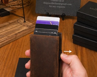 Puede incluir: Un tarjetero de cuero marrón con varias tarjetas visibles. El tarjetero se sostiene en una mano, con una caja negra en primer plano. El fondo incluye un letrero que dice "SHIPYARD - local leathersmith and engraving".