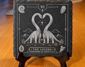 Könnte beinhalten: Ein quadratischer Untersetzer aus schwarzem Schiefer mit einer weißen Gravur im Tarotkarten-Stil mit der Aufschrift The Lovers. Das Motiv zeigt zwei langhalsige Dinosaurier, deren Hälse ein Herz bilden, umgeben von Fossilien, Bergen und einem Stern.