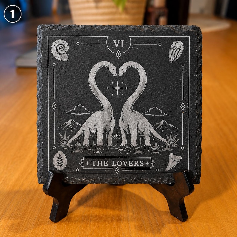 Puede incluir: Un posavasos cuadrado de pizarra negra con un grabado blanco de estilo carta del tarot titulado The Lovers. La ilustraci&oacute;n muestra dos dinosaurios de cuello largo con los cuellos curvados en forma de coraz&oacute;n, rodeados de f&oacute;siles, monta&ntilde;as y una estrella.