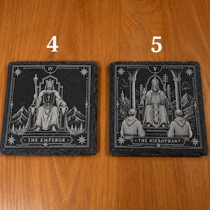 May include: Two square black slate coasters featuring white engraved tarot card designs. The left coaster shows The Emperor with the number IV, and the right coaster shows The Hierophant with the number V. Both feature detailed illustrations.