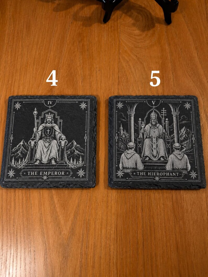 May include: Two square black slate coasters featuring white engraved tarot card designs. The left coaster shows The Emperor with the number IV, and the right coaster shows The Hierophant with the number V. Both feature detailed illustrations.