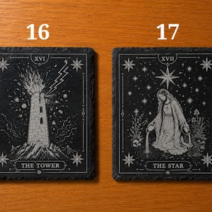 May include: Two rectangular black slate coasters featuring white engraved tarot card designs. The left coaster displays The Tower, labeled XVI, with a lightning bolt striking a stone tower. The right coaster shows The Star, labeled XVII, with a figure pouring water into a pool under a large star.