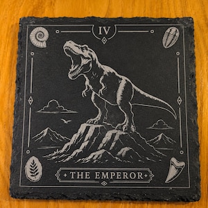 Puede incluir: Un posavasos cuadrado de pizarra negra con un grabado blanco de un Tyrannosaurus Rex sobre una cima monta&ntilde;osa. El dise&ntilde;o incluye el n&uacute;mero romano IV, el texto THE EMPEROR y peque&ntilde;os iconos de f&oacute;siles en las esquinas.