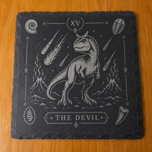 Puede incluir: Un posavasos cuadrado de pizarra negra con un dise&ntilde;o grabado con l&aacute;ser de un dinosaurio en el centro, rodeado de volcanes, meteoritos y f&oacute;siles. El n&uacute;mero romano XV est&aacute; en la parte superior y el texto THE DEVIL en la parte inferior.