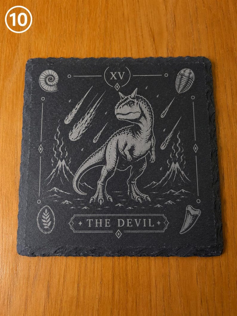 Puede incluir: Un posavasos cuadrado de pizarra negra con un dise&ntilde;o grabado con l&aacute;ser de un dinosaurio en el centro, rodeado de volcanes, meteoritos y f&oacute;siles. El n&uacute;mero romano XV est&aacute; en la parte superior y el texto THE DEVIL en la parte inferior.