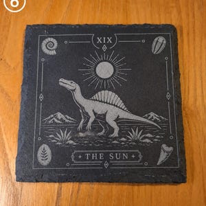 Puede incluir: Un posavasos cuadrado de pizarra negra con un Spinosaurus grabado con l&aacute;ser en el centro, un s&iacute;mbolo de sol arriba y el texto THE SUN en la parte inferior. El dise&ntilde;o incluye el n&uacute;mero romano XIX y cuatro iconos de f&oacute;siles en las esquinas.