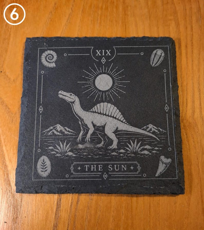 Puede incluir: Un posavasos cuadrado de pizarra negra con un Spinosaurus grabado con l&aacute;ser en el centro, un s&iacute;mbolo de sol arriba y el texto THE SUN en la parte inferior. El dise&ntilde;o incluye el n&uacute;mero romano XIX y cuatro iconos de f&oacute;siles en las esquinas.