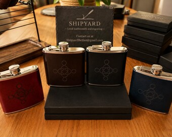 Könnte beinhalten: Edelstahl-Flachmänner in Burgunderrot, Braun, Schwarz und Marineblau, alle mit silbernen Verschlüssen und einem geometrischen Design. Auf einem Holzbrett steht "SHIPYARD - Local leathersmiths and engraving - Contact us at ShipyardBelfast@gmail.com".