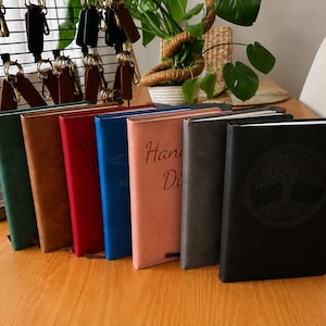 Könnte beinhalten: Eine Reihe von sieben rechteckigen Notizbüchern mit einfarbigen Einbänden in Grün, Braun, Rot, Blau, Rosa, Grau und Schwarz. Das schwarze Notizbuch zeigt ein kreisförmiges Baumdesign auf dem Einband, während das rosa Notizbuch den Text Hand Di trägt.