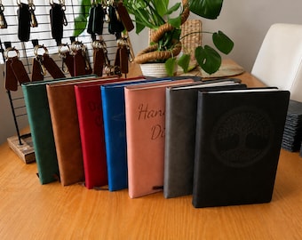 Könnte beinhalten: Eine Reihe von sieben rechteckigen Notizbüchern mit einfarbigen Einbänden in Grün, Braun, Rot, Blau, Rosa, Grau und Schwarz. Das schwarze Notizbuch zeigt ein kreisförmiges Baumdesign auf dem Einband, während das rosa Notizbuch den Text Hand Di trägt.