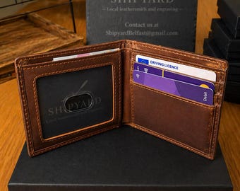 May include: Open brown leather wallet with visible card slots and a clear ID window. The wallet is displayed open, revealing cards and a driver's license. The wallet is on a black box, with a slate sign in the background that reads "SHIPYARD".