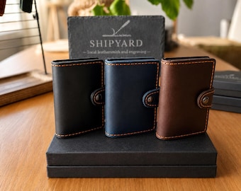 Könnte beinhalten: Drei Ledergeldbörsen in Schwarz, Marineblau und Braun, jeweils mit kontrastierenden orangefarbenen Nähten und einem Druckknopfverschluss. Die Geldbörsen werden auf einer schwarzen Box präsentiert, mit einem Schild "SHIPYARD - local leathersmith and engraving".