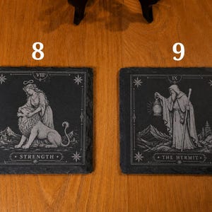 May include: Two square black slate coasters featuring white engraved tarot card designs. The left coaster displays the Strength card with a figure and lion, labeled VIII and STRENGTH. The right coaster shows The Hermit with a lantern, labeled IX and THE HERMIT.