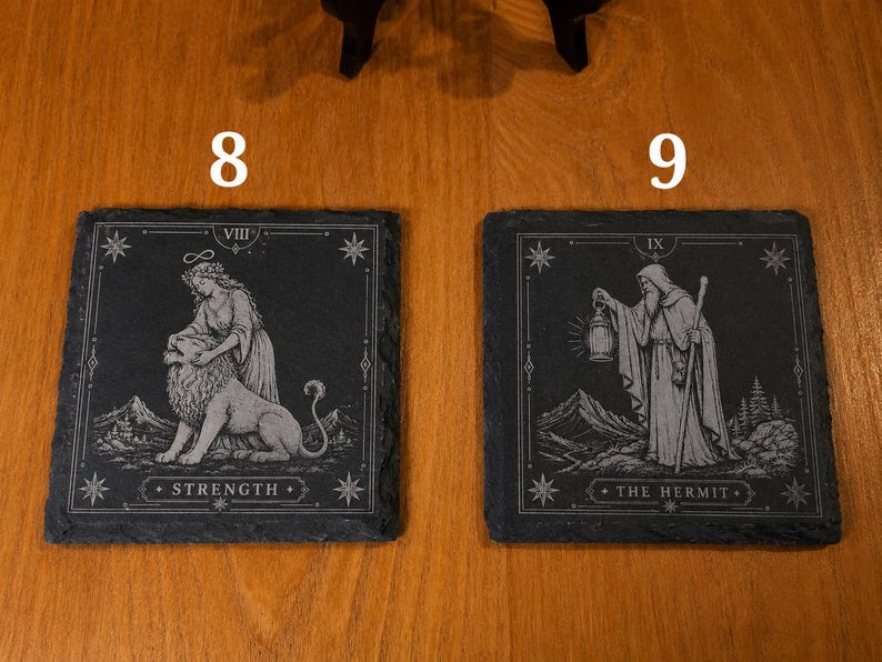 May include: Two square black slate coasters featuring white engraved tarot card designs. The left coaster displays the Strength card with a figure and lion, labeled VIII and STRENGTH. The right coaster shows The Hermit with a lantern, labeled IX and THE HERMIT.
