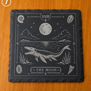 Puede incluir: Un posavasos cuadrado de pizarra negra con un grabado l&aacute;ser de un mosasaurio nadando en el agua bajo una luna llena. El dise&ntilde;o incluye el texto XVIII y THE MOON, rodeado de f&oacute;siles, nubes y estrellas con un estilo de carta de tarot.