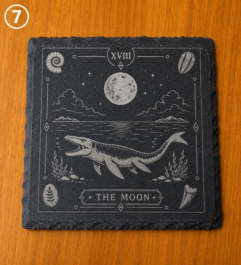 Puede incluir: Un posavasos cuadrado de pizarra negra con un grabado l&aacute;ser de un mosasaurio nadando en el agua bajo una luna llena. El dise&ntilde;o incluye el texto XVIII y THE MOON, rodeado de f&oacute;siles, nubes y estrellas con un estilo de carta de tarot.