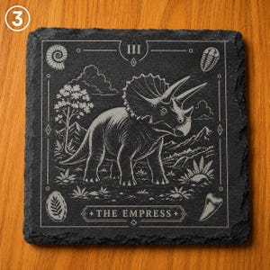 Puede incluir: Un posavasos cuadrado de pizarra negra con una ilustraci&oacute;n grabada a l&aacute;ser de un dinosaurio triceratops al estilo de una carta del tarot. El dise&ntilde;o incluye el n&uacute;mero romano III, el texto THE EMPRESS y varios f&oacute;siles prehist&oacute;ricos.