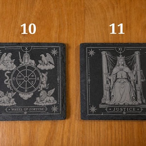 May include: Two square black slate coasters featuring engraved tarot card designs. The left coaster, labeled 10, depicts the Wheel of Fortune with celestial figures. The right coaster, labeled 11, shows the Justice card with a seated figure holding scales.