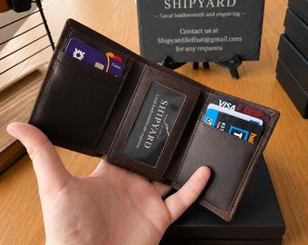 Puede incluir: Una cartera de cuero marrón oscuro de tres pliegues con múltiples ranuras para tarjetas y una ventana central transparente para identificación. La cartera está abierta, mostrando varias tarjetas de crédito y débito. El texto SHIPYARD es visible.