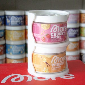 Może przedstawiać: Biały plastikowy uchwyt z dwoma słoikami produktów "More Chunky Flavour". Górny słoik jest różowy o smaku kruszonki truskawkowej, a dolny słoik jest pomarańczowy o smaku budyniu waniliowego. Inne słoiki są widoczne w tle.
