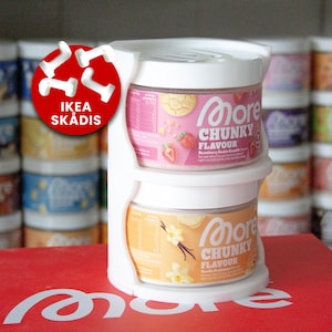 Może przedstawiać: Biały plastikowy uchwyt z dwoma słoikami "More Chunky Flavour". Górny słoik jest różowy o smaku "Strawberry Cookie Crumble", a dolny słoik jest pomarańczowy o smaku "Vanilla Perfection". Widoczne jest logo IKEA SKÅDIS.