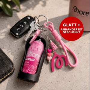 Könnte beinhalten: Ein Schlüsselanhänger mit einer rosa More Zerup Pink Boost Flasche in einer schwarzen Halterung, befestigt an einem silbernen Ring mit rosa geflochtenem Band, Quaste, einem rosa M-Anhänger und drei kleinen herzförmigen Perlen in Rosatönen.