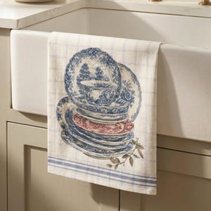 May include: A rectangular kitchen towel featuring a light cream background with a blue grid pattern and horizontal blue stripes at the bottom. The center displays a stack of blue and white patterned plates and a teacup with one red plate.