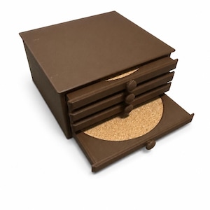 May include: A brown coaster set with a square holder. The holder has a drawer design with four coasters visible, one pulled out. The coasters are round with a cork surface. The set is made of a dark brown material.