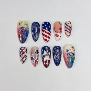 May include: A set of ten artificial nails with patriotic designs. The nails feature red, white, and blue colors, with patterns including stars, stripes, and fireworks. Some nails have glitter and small embellishments.