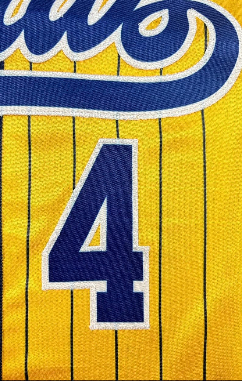 May include: A yellow athletic jersey featuring thin vertical black pinstripes and the large number 4 in dark blue with a white border. The upper portion displays a partial dark blue script logo with a white outline, all rendered in fabric.