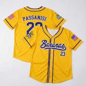 May include: A yellow baseball jersey with purple trim, featuring the text Bananas and the number 23 on the front. The back displays the name Passanisi and the number 23. Both sleeves feature a small American flag patch and a graphic logo.
