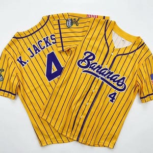 May include: A yellow baseball jersey with thin dark purple pinstripes, featuring the word Bananas and the number 4 on the front. The back displays K. JACKS and the number 4. It includes a small American flag patch on the sleeve and button closure.