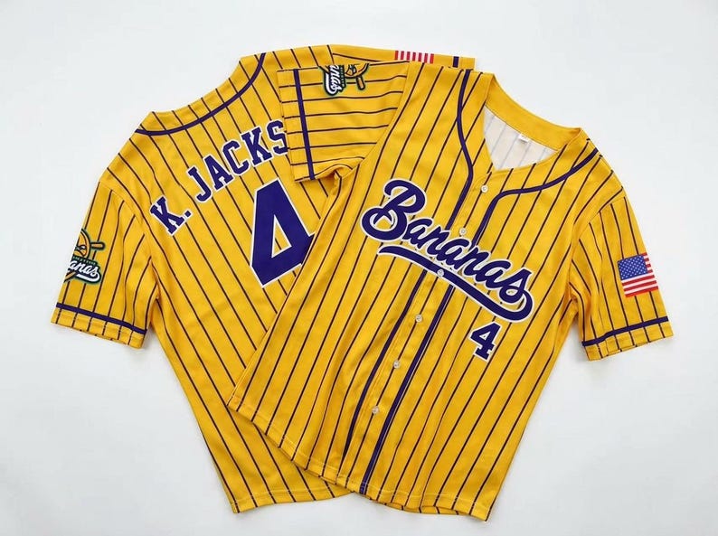May include: A yellow baseball jersey with thin dark purple pinstripes, featuring the word Bananas and the number 4 on the front. The back displays K. JACKS and the number 4. It includes a small American flag patch on the sleeve and button closure.