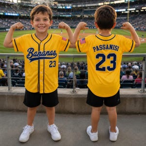 May include: A yellow baseball jersey with navy blue trim, featuring the word Bananas in script, the number 23, and a small American flag patch on the sleeve. The back displays the name Passanisi above the number 23 in navy blue lettering.