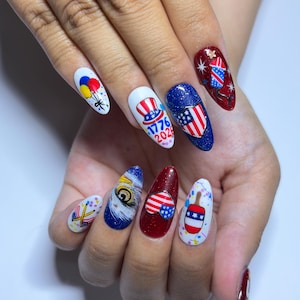 May include: A set of almond-shaped, painted fingernails with patriotic designs. The nails feature balloons, an eagle eye, the American flag, and the text "1776 2023". Colors include red, white, blue, yellow, and gold.