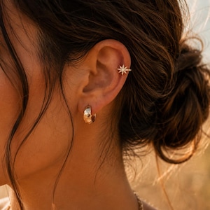 Peut inclure: Une créole dorée à la surface texturée et un petit bijou d'oreille en forme d'étoile avec des pierres claires incrustées. Le motif en étoile présente de multiples pointes rayonnantes et est placé sur le cartilage supérieur de l'oreille.