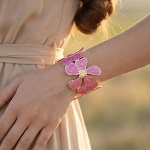 Könnte beinhalten: Ein goldfarbenes Armband mit einem floralen Design und rosa Emaille-Blütenblättern. Das Armband wird am Handgelenk getragen und zeigt die komplizierten Details der blütenförmigen Verzierungen. Der Hintergrund ist unscharf und hebt den Schmuck hervor.