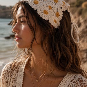 Puede incluir: Un pañuelo de crochet color crema con detalles de margaritas amarillas. El pañuelo está atado en la parte trasera. La imagen también muestra un top de crochet color crema y collares dorados. Un accesorio de moda.
