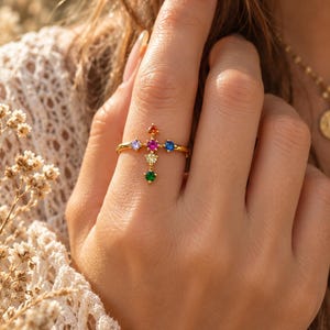 Könnte beinhalten: Ein goldfarbener Ring mit einer kreuzförmigen Anordnung aus sechs kleinen, runden, bunten Edelsteinen. Die Steine umfassen einen roten Stein oben, einen violetten links, einen blauen rechts, einen pinken in der Mitte, einen gelben unten und einen grünen am unteren Ende.