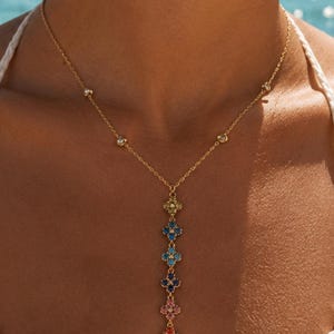 Op de afbeelding: Een goudkleurige lariat-ketting met een fijne ketting met kleine ingezette heldere steentjes en een verticale hanger bestaande uit zes veelkleurige bloemvormige bedels in geel, lichtblauw, donkerblauw, roze, oranje en rood.