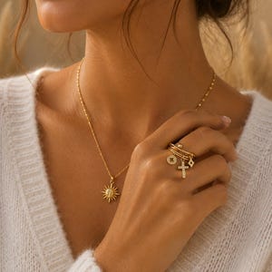 Peut inclure: Un collier en or avec un pendentif soleil et une chaîne délicate. L'image montre également une main portant plusieurs bagues en or, dont une bague croix et une bague à breloques. La personne porte un pull blanc.