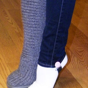 May include: A pair of gray knitted socks and a pair of white socks with a black strap and a pink button. The socks are worn by a person standing on a wooden floor.