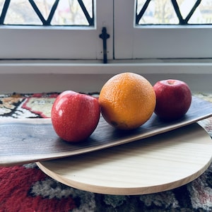 May include: A wooden serving tray with a dark gray, rectangular top and a light wood, circular base. The tray holds a bright orange, a red apple, and another red apple. The tray is on a patterned rug near a window.