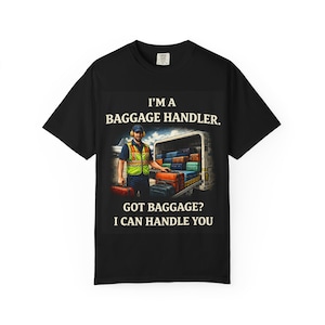 Puede incluir: Camiseta negra con el texto "I'M A BAGGAGE HANDLER. GOT BAGGAGE? I CAN HANDLE YOU." El gráfico muestra un manipulador de equipaje con equipaje en un camión.