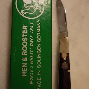 May include: A pocket knife with a silver blade and a dark brown handle, next to a green box. The box has the words "HEN & ROOSTER" and "WORLD'S FINEST" printed on it. The box also says "MADE IN SOLINGEN, GERMANY."