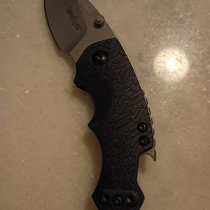 May include: A black and silver folding knife with a curved blade. The knife has a textured black handle and a silver blade with the brand name "Kershaw" printed on it. The knife also has a small metal loop at the base of the handle.